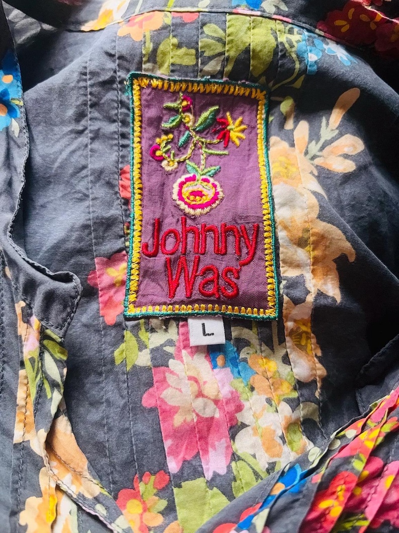 Puede incluir: Primer plano de una camisa con estampado floral y un parche decorativo. El parche es morado con flores bordadas y el texto "Johnny Was" en rojo. La camisa es gris con un estampado floral colorido y una etiqueta de talla "L".