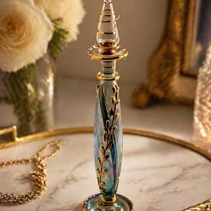 Può includere: Un'alta e ornata bottiglia di profumo in vetro con un design blu e oro. La bottiglia presenta un corpo sottile e allungato con accenti di foglie d'oro e un tappo decorativo e appuntito. La base e le finiture sono dorate.