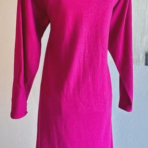 May include: A long-sleeved, knee-length dress in a vibrant magenta color. The garment features a high, straight boat neckline and a simple, straight-cut silhouette. The fabric appears smooth and solid, with long sleeves extending to the wrists.