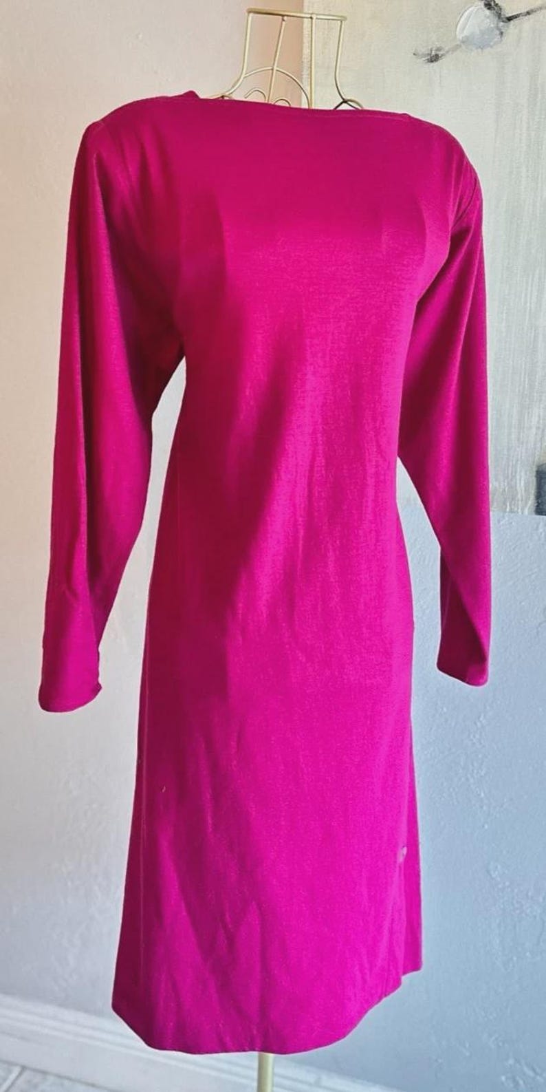 May include: A long-sleeved, knee-length dress in a vibrant magenta color. The garment features a high, straight boat neckline and a simple, straight-cut silhouette. The fabric appears smooth and solid, with long sleeves extending to the wrists.
