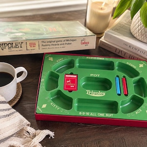May include: A green Tripoley game board with a red box, a blue peg, and a red peg. The board features printed text including Pot, Kitty, Ace, King-Queen, and 8-9-10 All One Suit. The game board is rectangular with molded compartments for play.