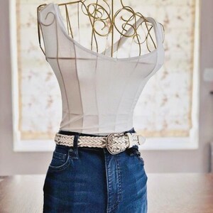 May include: A decorative dress form with a gold wire frame, a white tank top, and dark blue denim jeans. A white braided belt with a decorative buckle is around the waist. The form is displayed on a wooden table.