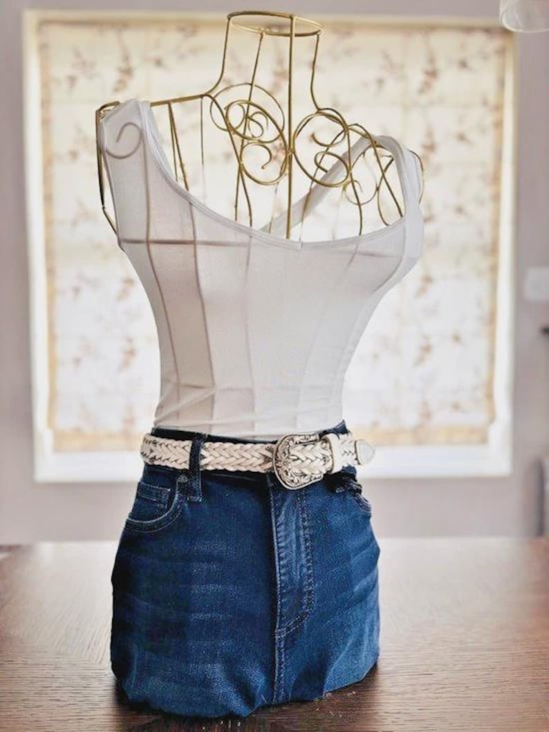May include: A decorative dress form with a gold wire frame, a white tank top, and dark blue denim jeans. A white braided belt with a decorative buckle is around the waist. The form is displayed on a wooden table.
