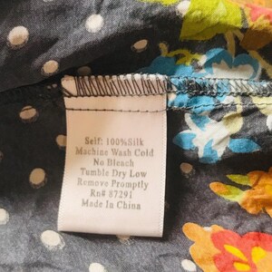 Puede incluir: Primer plano de una tela de seda con un estampado floral en naranja, azul y verde sobre un fondo gris oscuro con lunares blancos. Una etiqueta dice "Self: 100% Silk, Machine Wash Cold, No Bleach, Tumble Dry Low, Remove Promptly, Rn# 87291, Made In China."