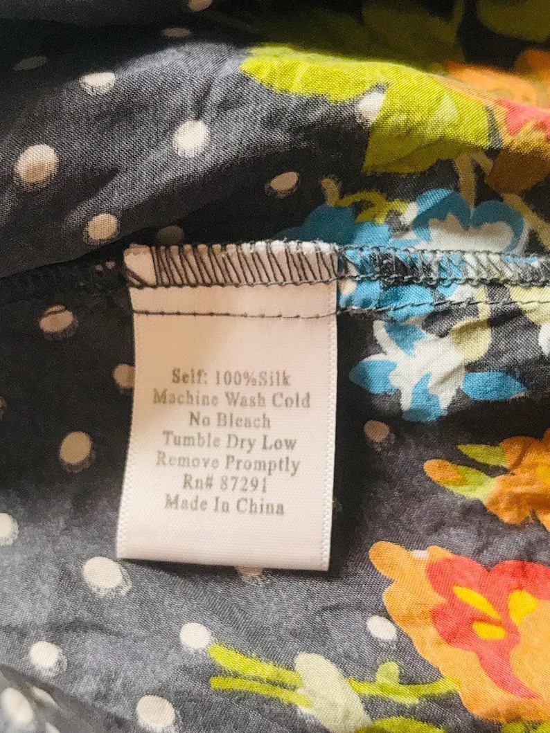 Puede incluir: Primer plano de una tela de seda con un estampado floral en naranja, azul y verde sobre un fondo gris oscuro con lunares blancos. Una etiqueta dice "Self: 100% Silk, Machine Wash Cold, No Bleach, Tumble Dry Low, Remove Promptly, Rn# 87291, Made In China."