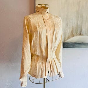 May include: A long-sleeved, cream-colored blouse featuring a subtle, tonal abstract pattern. The garment has a structured collar, a button-down front placket, and a fitted waist section that transitions into a pleated peplum hemline.