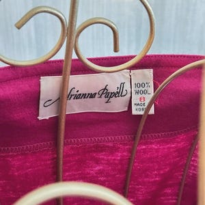 May include: A magenta garment featuring a white interior label with the text Adrianna Papell. A smaller adjacent white tag displays 100% WOOL, the size 8, and MADE IN KOREA. The fabric has a visible textured weave and is positioned behind a gold wire stand.