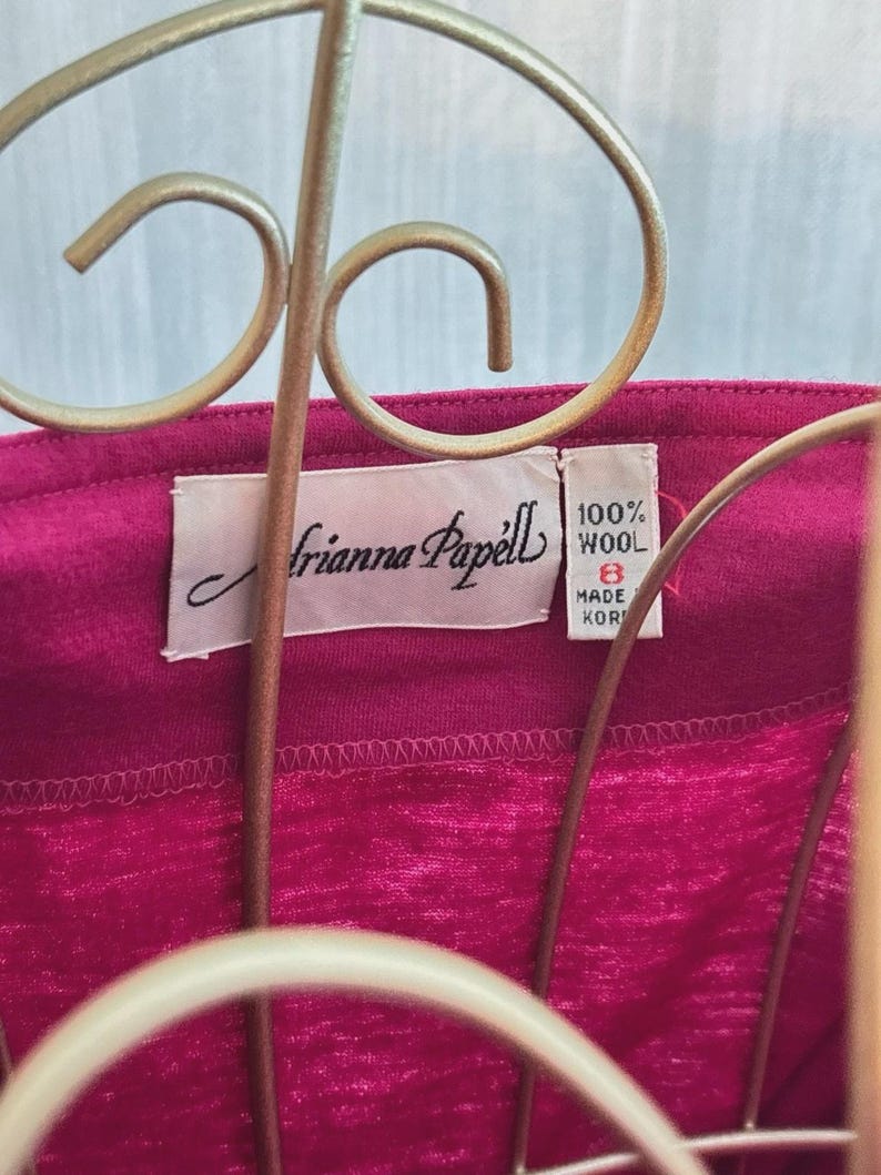 May include: A magenta garment featuring a white interior label with the text Adrianna Papell. A smaller adjacent white tag displays 100% WOOL, the size 8, and MADE IN KOREA. The fabric has a visible textured weave and is positioned behind a gold wire stand.