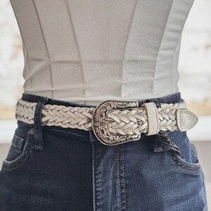 May include: Cream-colored braided belt with a decorative silver-tone buckle. The buckle features a floral design. The belt is worn with dark blue denim jeans and a white top. A stylish accessory for any outfit.