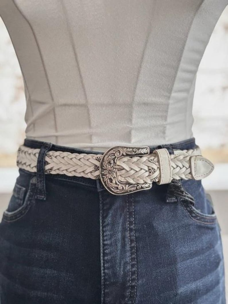 May include: Cream-colored braided belt with a decorative silver-tone buckle. The buckle features a floral design. The belt is worn with dark blue denim jeans and a white top. A stylish accessory for any outfit.