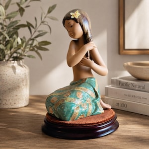 Può includere: Una statuetta in porcellana raffigurante una persona con lunghi capelli castani adornati da un piccolo fiore giallo, inginocchiata e che indossa un pareo con motivi turchesi e arancioni. La figura è montata su una base circolare in legno.