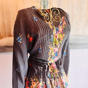 Puede incluir: Un vestido gris oscuro con mangas largas y cuello en V, adornado con pliegues y bordados florales. El vestido presenta una cintura con cord&oacute;n y una falda con un vibrante estampado floral en tonos rosa, azul y amarillo.