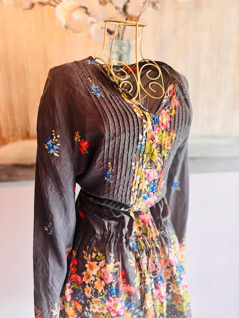 Puede incluir: Un vestido gris oscuro con mangas largas y cuello en V, adornado con pliegues y bordados florales. El vestido presenta una cintura con cord&oacute;n y una falda con un vibrante estampado floral en tonos rosa, azul y amarillo.