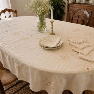May include: An oval-shaped cream-colored tablecloth featuring intricate white floral embroidery along the edges and center. The fabric is draped over a table, accompanied by a stack of matching embroidered napkins on a plate and a glass vase.