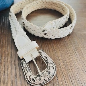 May include: A white braided belt with a silver-colored, ornate buckle. The belt is made of woven material and features a classic western design. The buckle has a floral pattern.