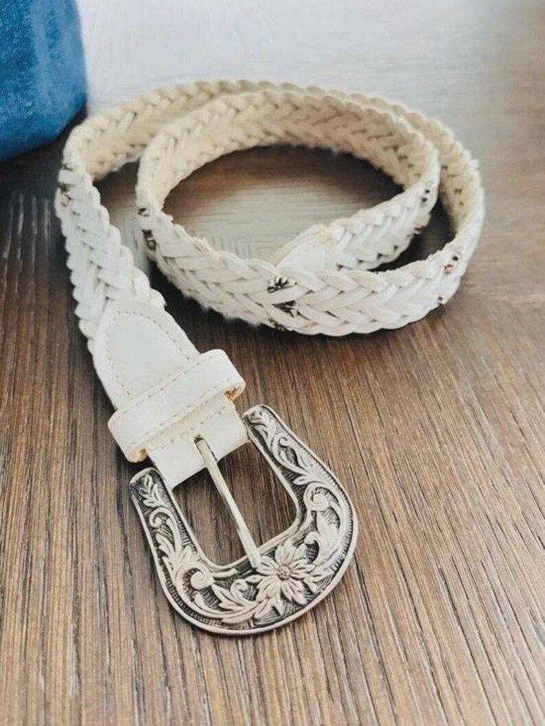 May include: A white braided belt with a silver-colored, ornate buckle. The belt is made of woven material and features a classic western design. The buckle has a floral pattern.