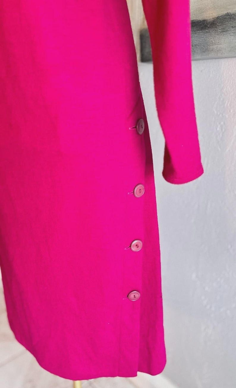 May include: A vibrant fuchsia long-sleeved garment featuring a vertical row of four matching circular buttons aligned along the side seam. The fabric has a smooth, solid texture, and the design includes a simple, clean silhouette with long sleeves.