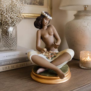 May include: A porcelain figurine of a person with dark hair and a white floral crown, sitting cross-legged on a circular green base. The figure wears a white cloth draped over the lap, holding a cluster of small flowers, set on a gold-toned stand.