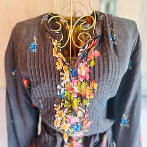 Puede incluir: Una blusa gris oscuro con mangas largas y cuello en V, adornada con pliegues verticales y un estampado floral. El dise&ntilde;o floral presenta una mezcla de rosa, azul, amarillo y verde. La blusa se exhibe en un maniqu&iacute; dorado.