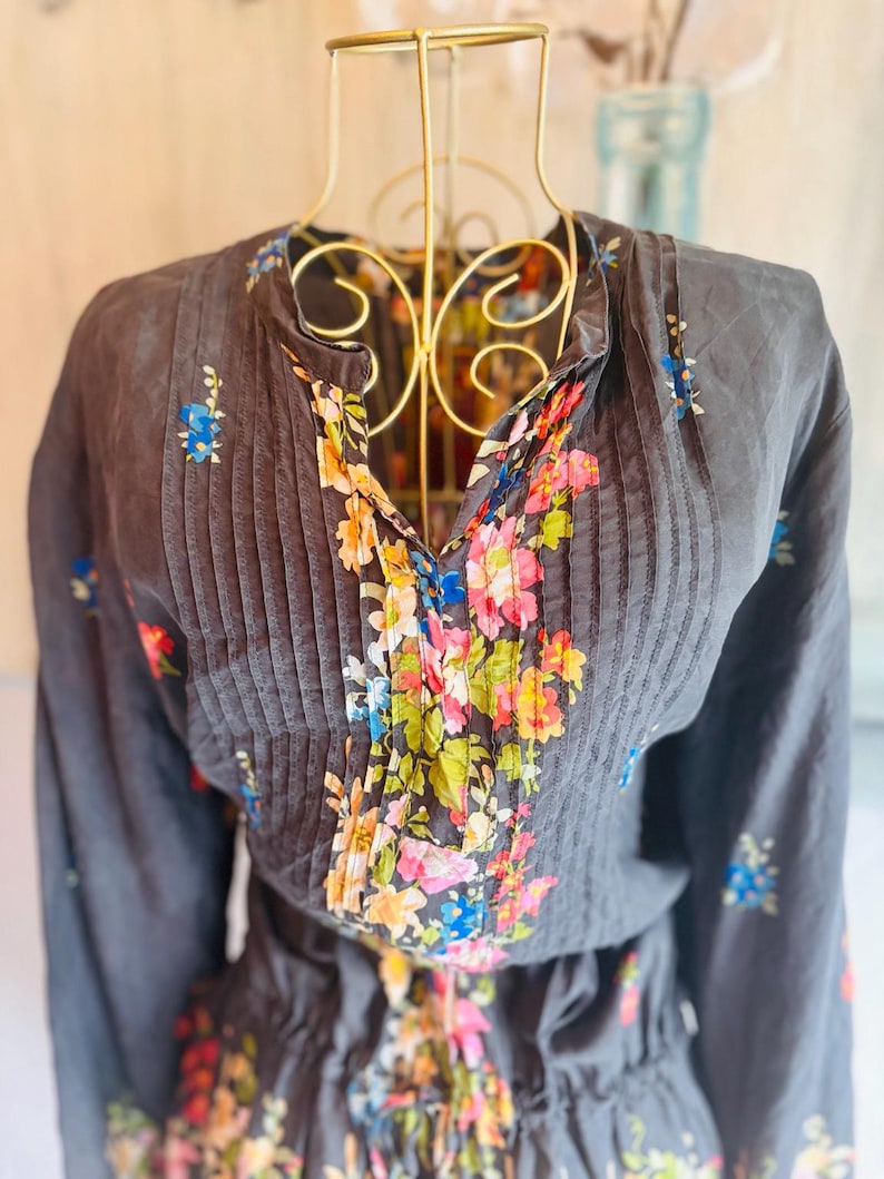 Puede incluir: Una blusa gris oscuro con mangas largas y cuello en V, adornada con pliegues verticales y un estampado floral. El dise&ntilde;o floral presenta una mezcla de rosa, azul, amarillo y verde. La blusa se exhibe en un maniqu&iacute; dorado.