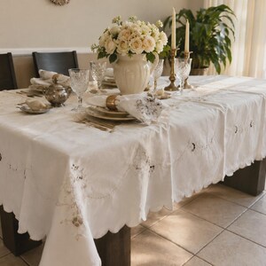 Pode incluir: Uma toalha de mesa retangular branca com bordados intrincados ao longo das bordas recortadas. O tecido apresenta delicados padrões florais e detalhes de costura, disposta sobre uma mesa de jantar com pratos, copos e um vaso de flores.