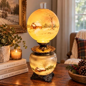 May include: A hurricane lamp featuring a spherical glass shade and a rounded glass base, both decorated with a painted winter landscape of snow-covered cabins and bare trees. The lamp has a dark metal base and a clear glass chimney at the top.