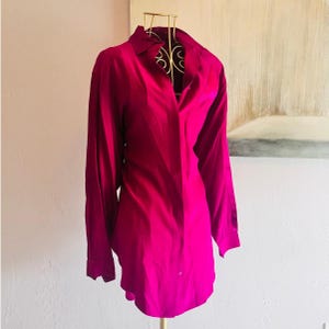 May include: A long-sleeved button-down shirt in a vibrant magenta color. The shirt features a classic collar, a concealed front placket, and long sleeves with cuffs. The fabric has a smooth, slightly reflective finish, displayed on a wire mannequin.