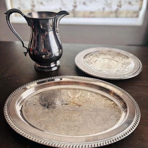 Op de afbeelding: Een zilverkleurige kan met een gebogen handvat en een kleine tuit, samen met twee zilveren serveerschalen. De schalen hebben sierlijke bloempatronen en decoratieve randen. De items zijn geplaatst op een donker houten oppervlak.