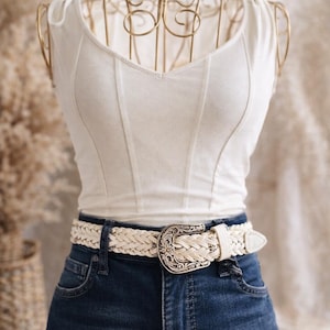 May include: A white corset-style top paired with dark blue denim jeans and a braided white belt with a decorative silver buckle. The top has a V-neck and vertical seams.
