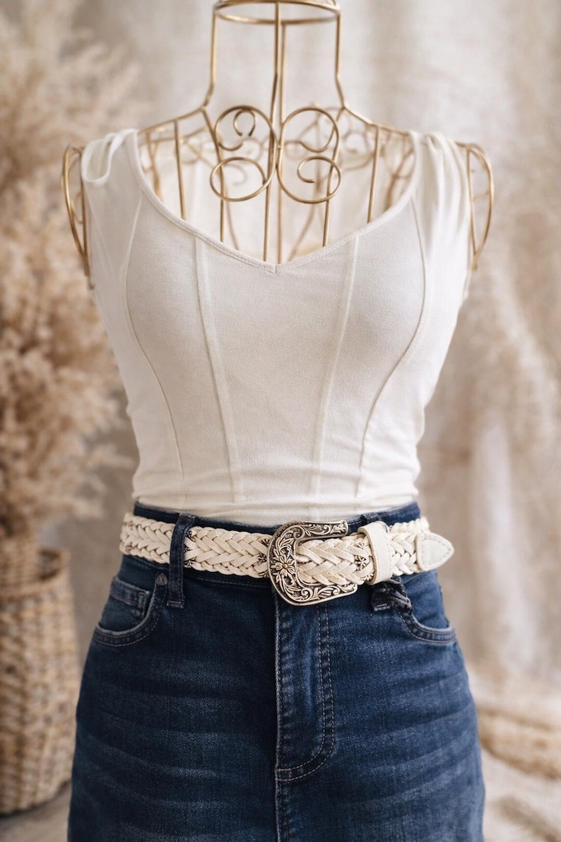 May include: A white corset-style top paired with dark blue denim jeans and a braided white belt with a decorative silver buckle. The top has a V-neck and vertical seams.