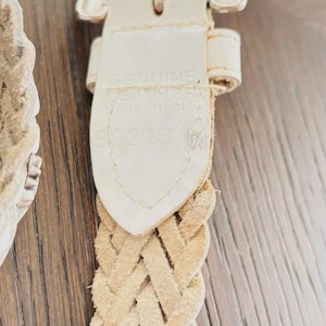 May include: Close-up of a cream-colored braided leather belt with a silver-toned buckle. The belt's tag reads "GENUINE LEATHER MADE IN INDIA" and includes the number "96218". The belt is on a wooden surface.