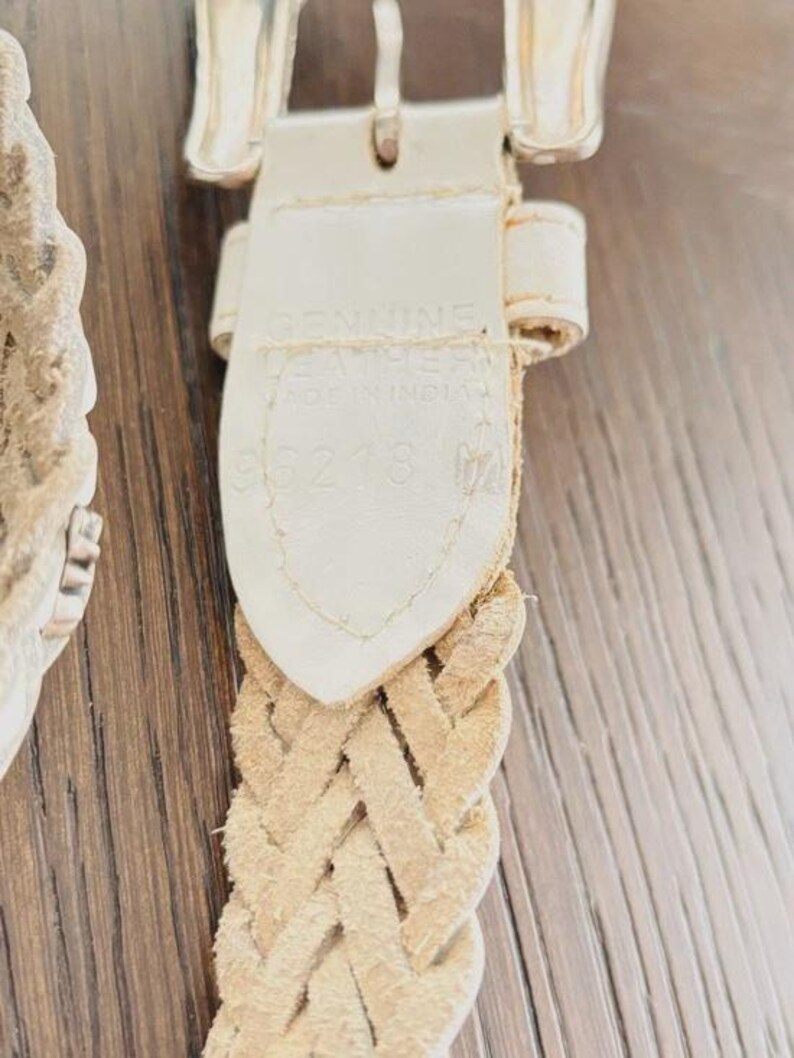 May include: Close-up of a cream-colored braided leather belt with a silver-toned buckle. The belt's tag reads "GENUINE LEATHER MADE IN INDIA" and includes the number "96218". The belt is on a wooden surface.