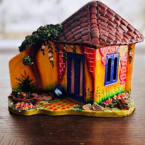 May include: A colorful miniature house with a brown tiled roof, blue door and window frames, and a yellow and orange facade. The house is decorated with brick accents, flowers, and a small tree.