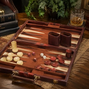 May include: A wooden backgammon set featuring a dark brown folding case with brass latches, a felt playing surface, cream and dark brown triangular points, cream and dark brown checkers, two leather dice cups, and several dice in red and white.