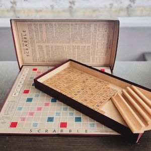 Pode incluir: Um jogo de tabuleiro Scrabble vintage com um tabuleiro dobrável com uma grelha de quadrados coloridos, uma tampa de caixa escura com regras impressas, um tabuleiro com peças de letras de madeira e quatro suportes de madeira.