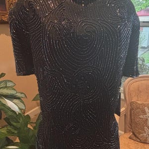 May include: Black beaded top with short sleeves and a high neckline. The top is covered in intricate swirling patterns of beads, creating a textured and shimmering effect. The design is elegant and suitable for formal occasions.