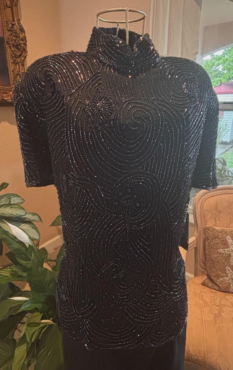 May include: Black beaded top with short sleeves and a high neckline. The top is covered in intricate swirling patterns of beads, creating a textured and shimmering effect. The design is elegant and suitable for formal occasions.