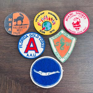 May include: Six embroidered fabric patches featuring various designs: a black cat with 'HSA HALLOWEEN MEET 1969', a globe with flags and 'USC&GS SHIP DISCOVERER SCOR 1970', a Santa Claus with 'MIAMI SHORES COUNTRY CLUB', 'FLORIDA GOLD COAST A.A.U.', a seahorse with 'SHEELER WINTON SWIM CLUB', and a swimmer.