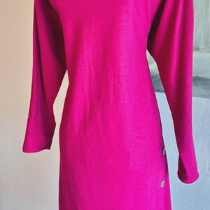 May include: A long-sleeved magenta dress featuring a boat neckline and a vertical row of four round buttons positioned along the lower left side. The garment is displayed on a gold-toned wire mannequin form, showcasing a solid, vibrant color.