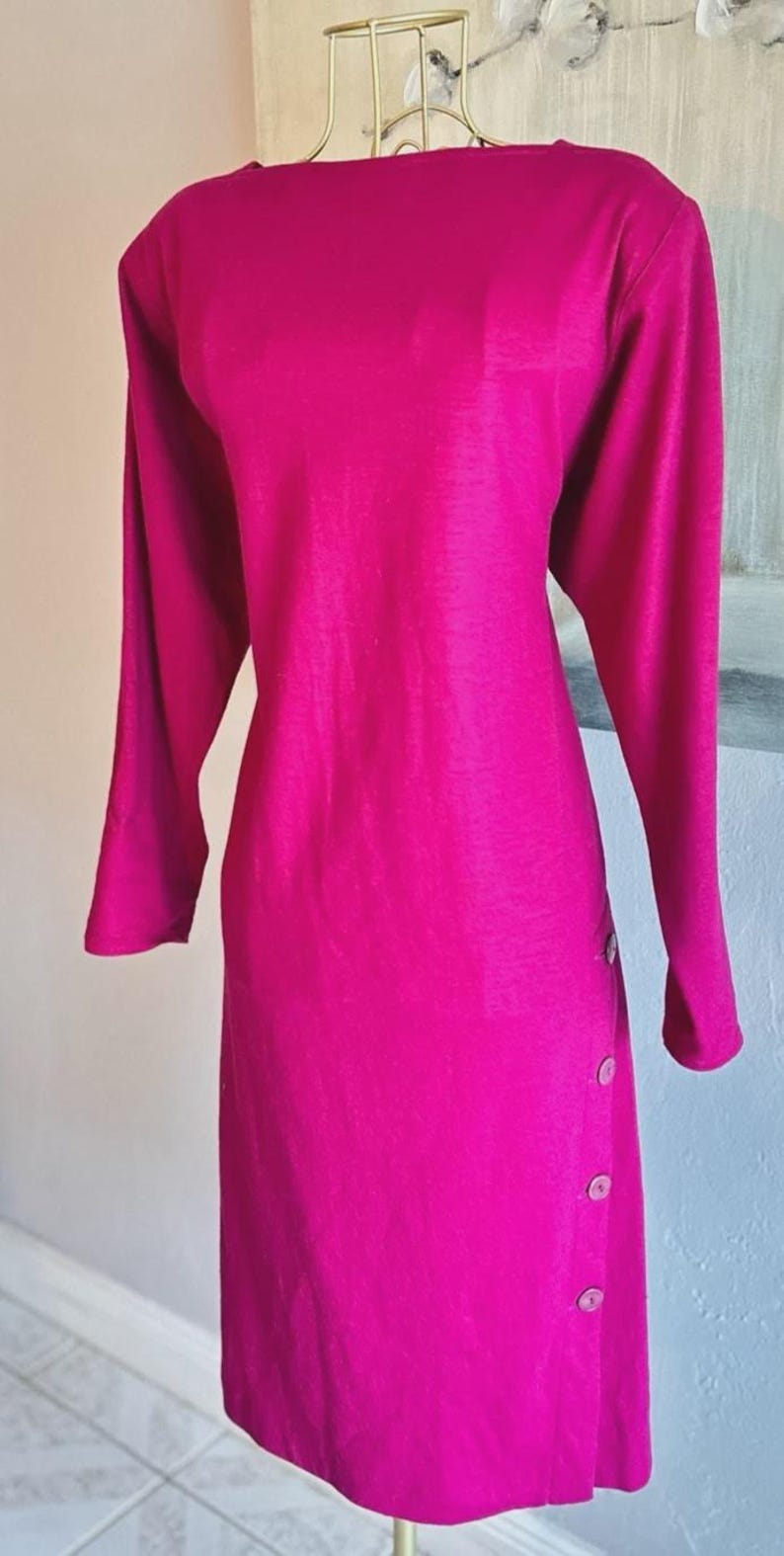 May include: A long-sleeved magenta dress featuring a boat neckline and a vertical row of four round buttons positioned along the lower left side. The garment is displayed on a gold-toned wire mannequin form, showcasing a solid, vibrant color.