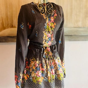 Puede incluir: Vestido marr&oacute;n oscuro con mangas largas y cuello en V, con estampado floral. El corpi&ntilde;o tiene pliegues verticales y la falda tiene un estampado de lunares en el dobladillo. El vestido se exhibe en un maniqu&iacute; dorado.