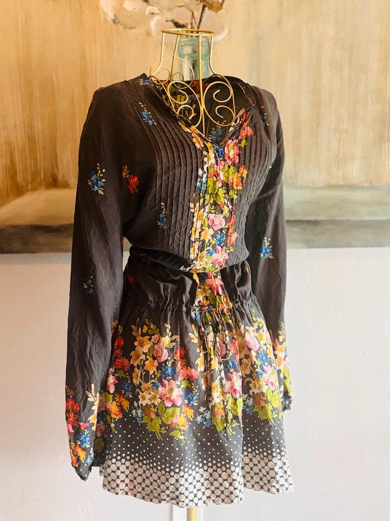 Puede incluir: Vestido marr&oacute;n oscuro con mangas largas y cuello en V, con estampado floral. El corpi&ntilde;o tiene pliegues verticales y la falda tiene un estampado de lunares en el dobladillo. El vestido se exhibe en un maniqu&iacute; dorado.