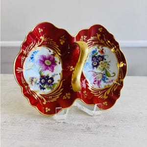 May include: A double-bowl porcelain serving dish featuring a deep red glaze with ornate gold filigree accents. The two white interior basins are decorated with colorful floral bouquets. A central gold handle connects the two scalloped sections.