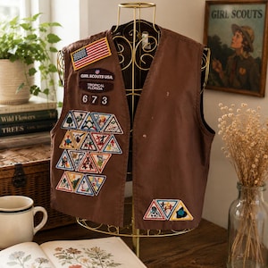 May include: A brown Girl Scouts vest featuring a United States flag patch, a Girl Scouts USA Tropical Florida patch, the number 673, and numerous triangular embroidered merit badges arranged on the front panels of the garment.