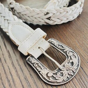 May include: White braided belt with a decorative silver buckle. The buckle features an ornate floral design. The belt is made of a woven material and has a matching white leather loop.