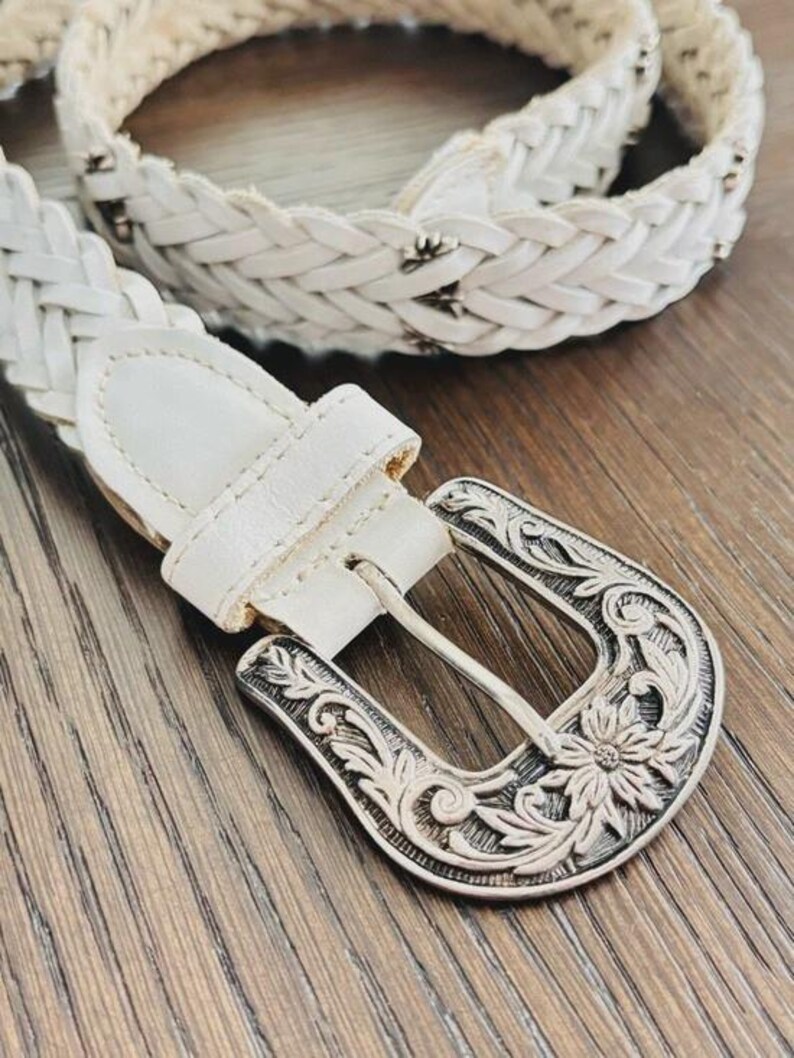 May include: White braided belt with a decorative silver buckle. The buckle features an ornate floral design. The belt is made of a woven material and has a matching white leather loop.