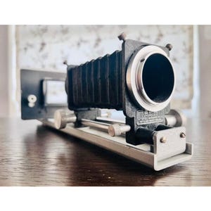 May include: A black bellows camera attachment featuring a silver circular lens mount, metal adjustment knobs, and a rectangular back plate. The device is mounted on a silver metal rail system with visible screws and a B.P.M. product label.
