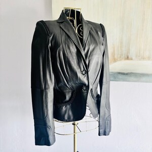 May include: A black leather blazer featuring a notched lapel, two front buttons, and decorative braided trim along the shoulders and pocket edges. The jacket has structured shoulders, long sleeves, and a tailored fit with visible panel seams.