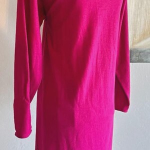 May include: A long-sleeved dress in a vibrant magenta color. The garment features a high, rounded neckline and a straight, knee-length silhouette. The fabric has a smooth, solid texture with long sleeves that extend to the wrists, displayed on a mannequin.