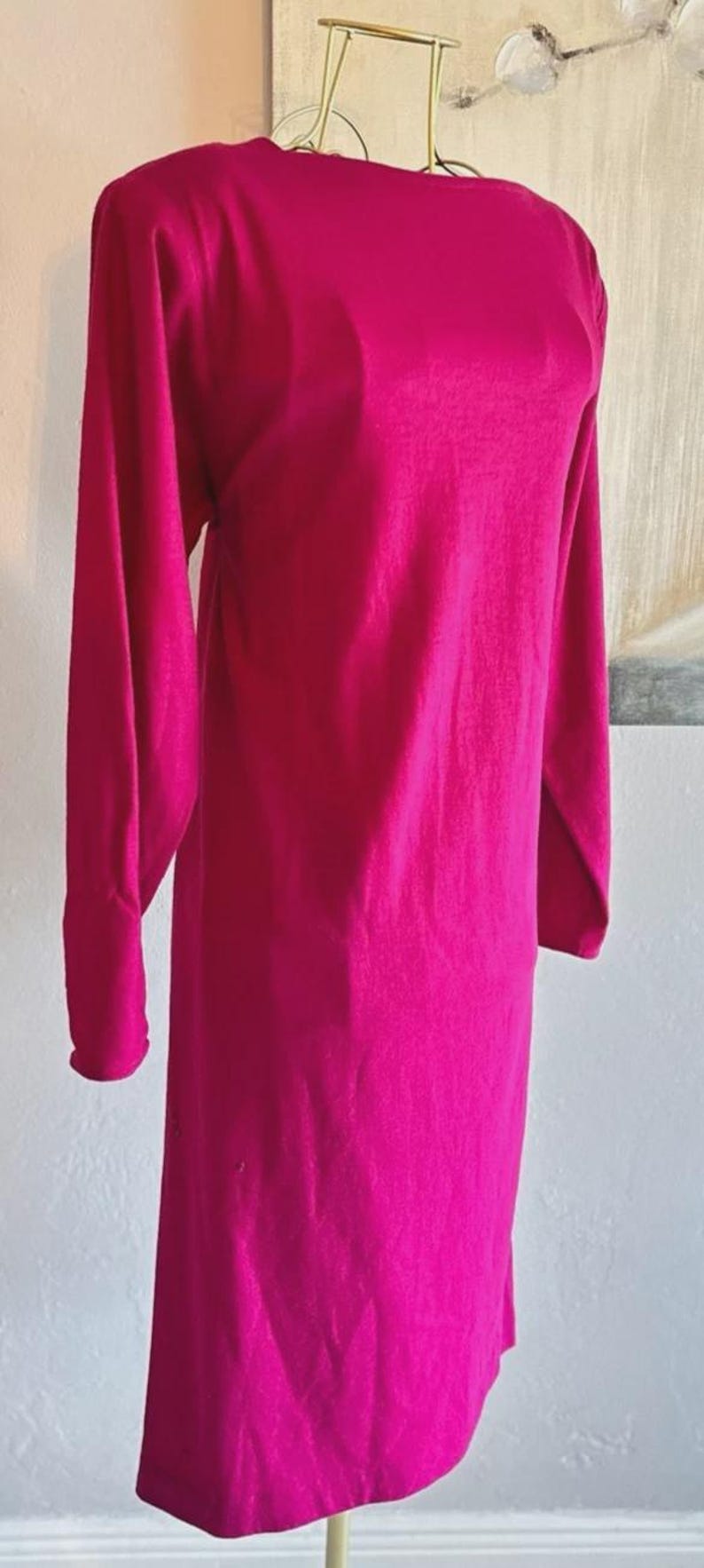 May include: A long-sleeved dress in a vibrant magenta color. The garment features a high, rounded neckline and a straight, knee-length silhouette. The fabric has a smooth, solid texture with long sleeves that extend to the wrists, displayed on a mannequin.